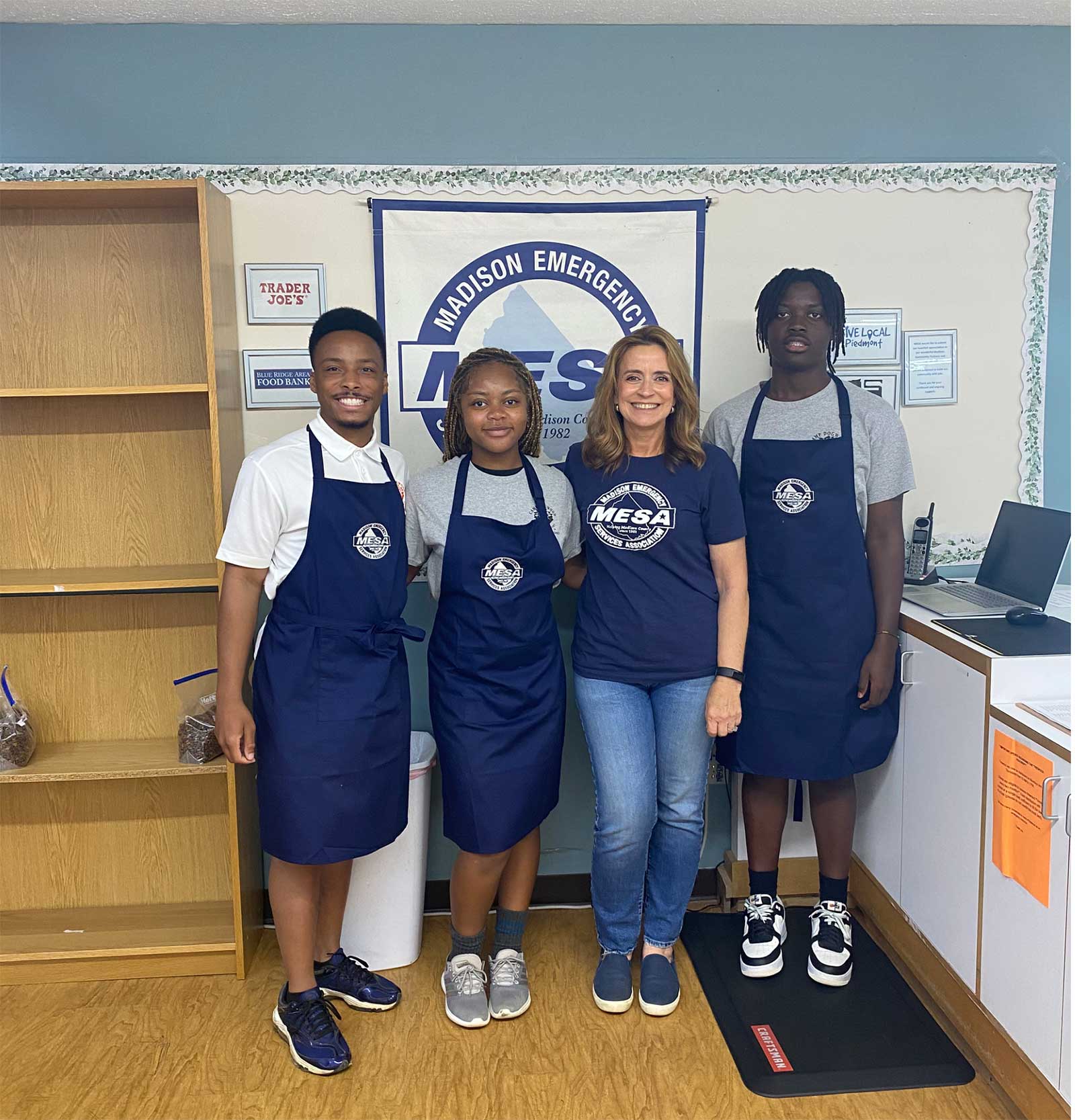 Four people standing indoors in front of a Madison Emergency Services Association banner, three wearing blue MESA aprons and one in a MESA t-shirt, smiling at the camera.