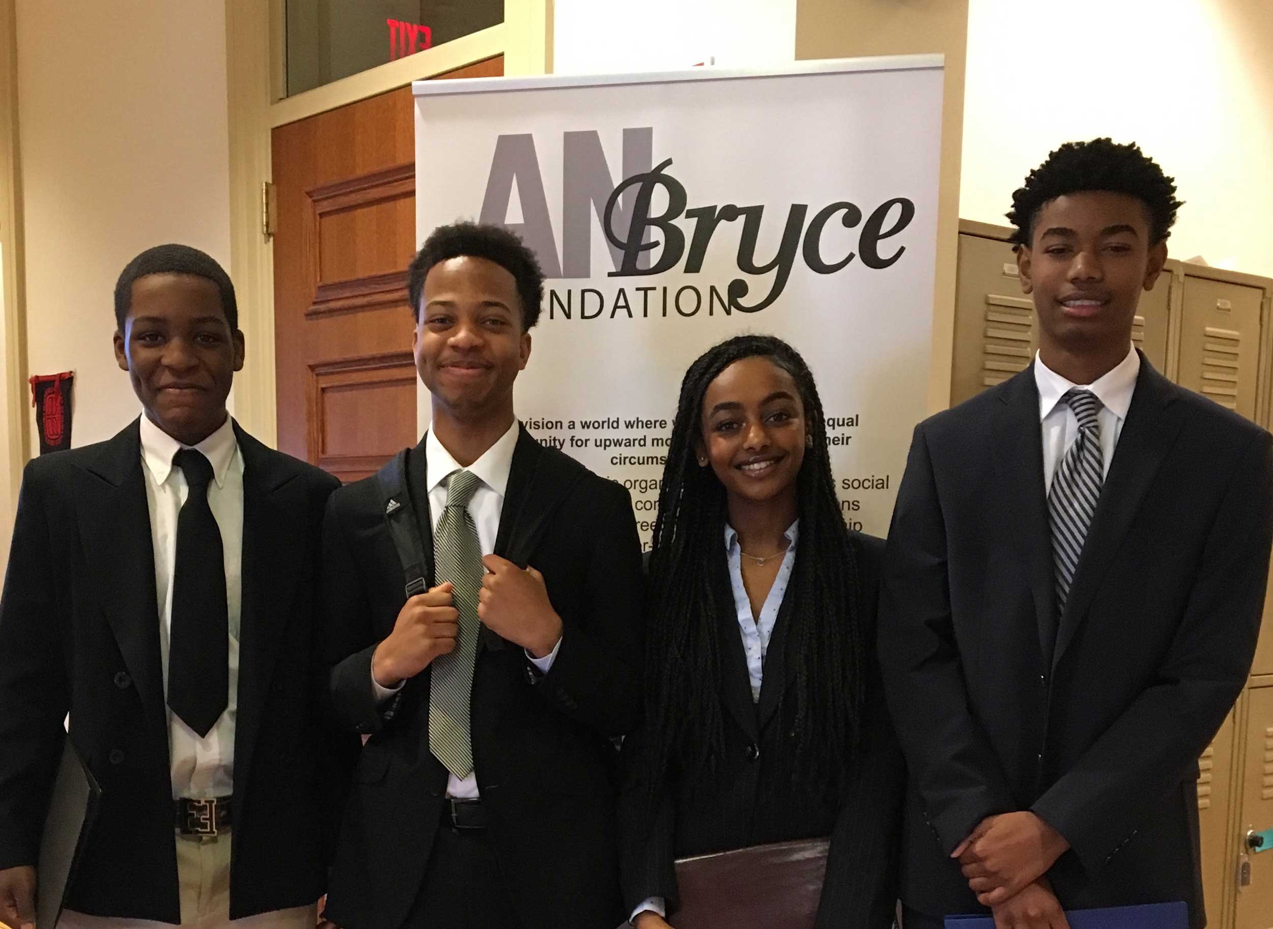 Four young individuals in the Pillors of Excellence Intership program dressed in formal attire standing in front of a Bryce Foundation banner inside a building with lockers.