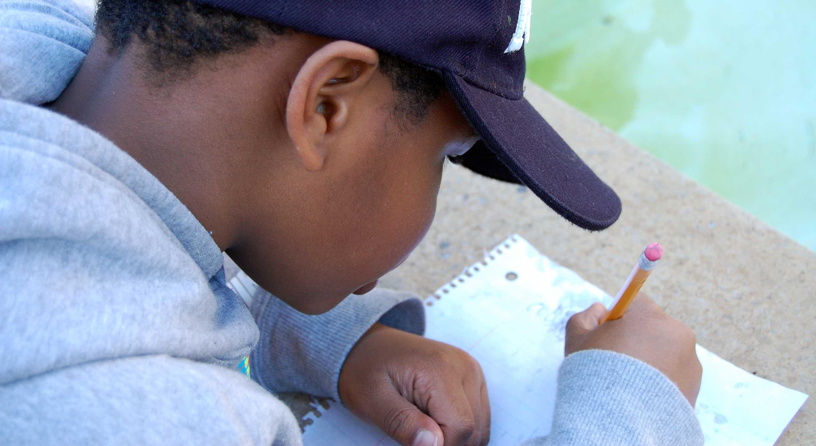 Young boy at Saturday Institute wearing a black cap and gray sweatshirt writing with a pencil on lined notebook paper outdoors.