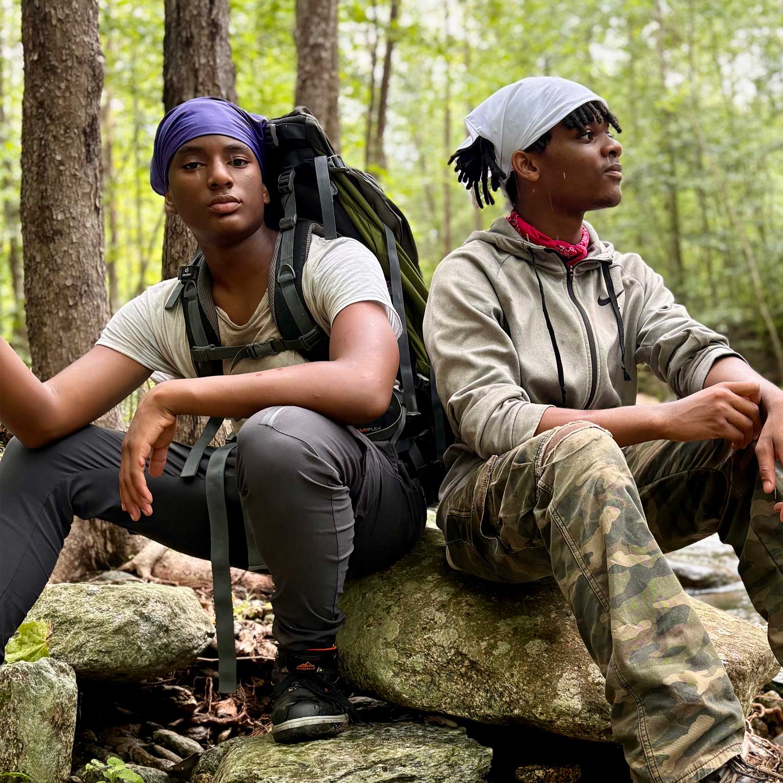 Two young men wearing headscarves sitting on rocks in a forested area of Camp Dogwood, one with a backpack and the other in camouflage pants.