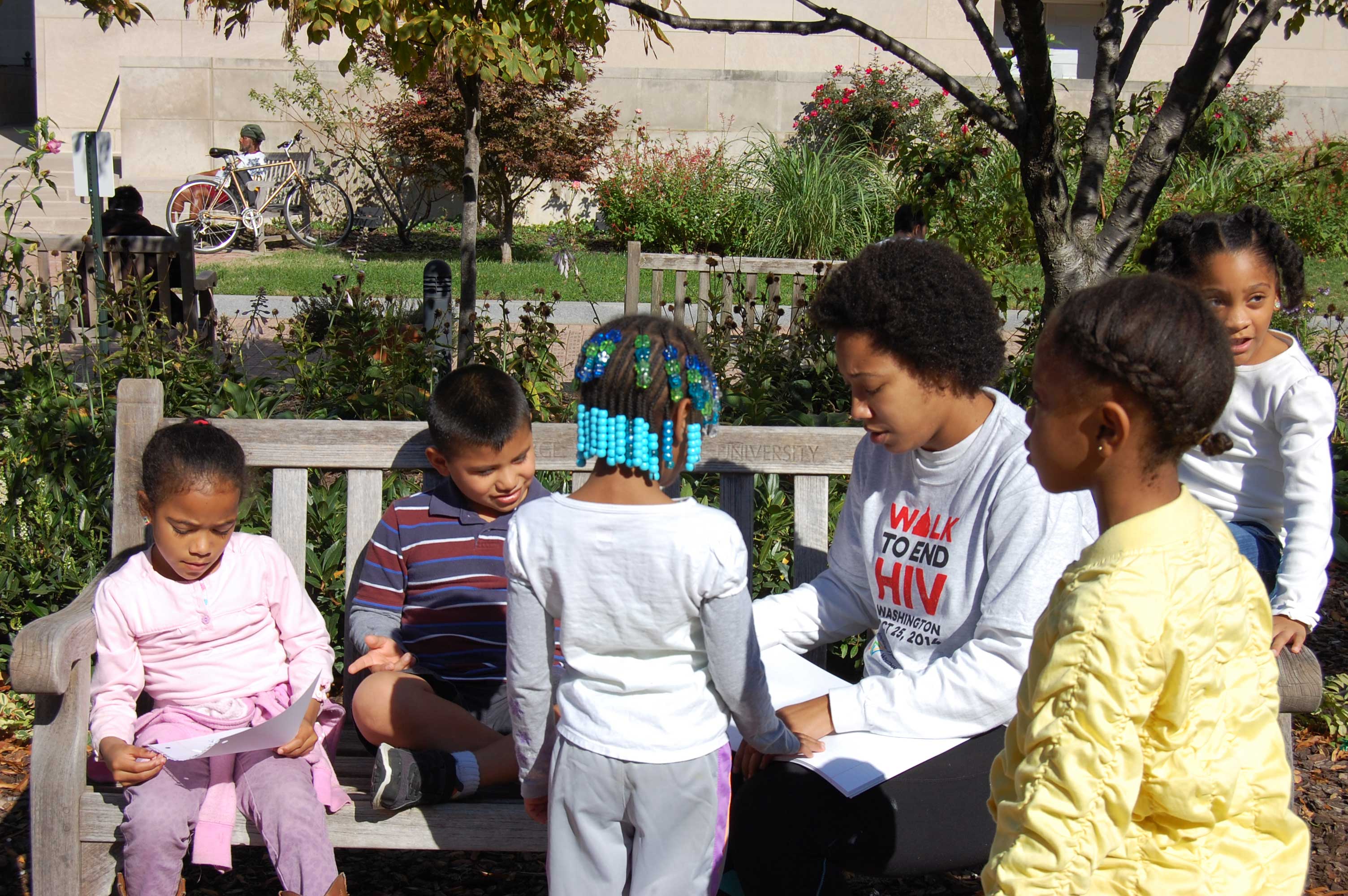 A group of children and a young adult at Saturday Institute sitting and standing around a wooden bench in a garden, with the young adult wearing a 'Walk to End HIV' shirt.