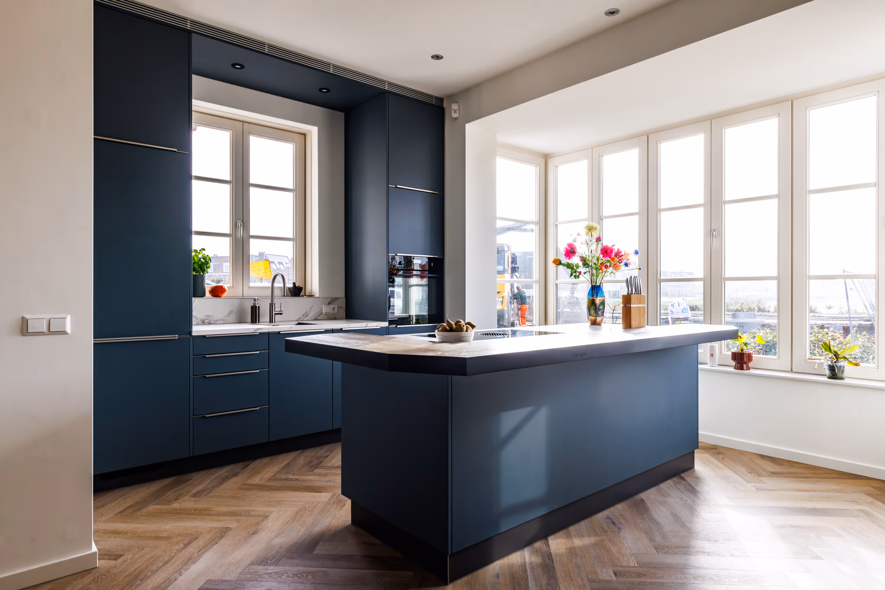 Moderne keuken met donkerblauwe kasten, centraal kookeiland, houten visgraatvloer en grote ramen met planten en een vaas met bloemen.