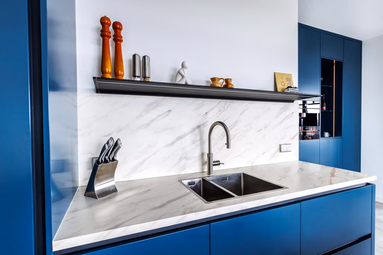 Moderne keuken met dubbele roestvrijstalen spoelbak, blauwe kasten, marmeren achterwand en keukenaccessoires op een plank.