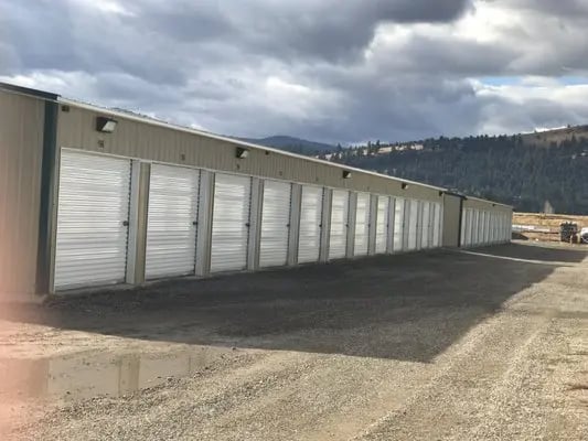 Long row of white storage unit doors on a gravel lot with cloudy sky and distant hills.