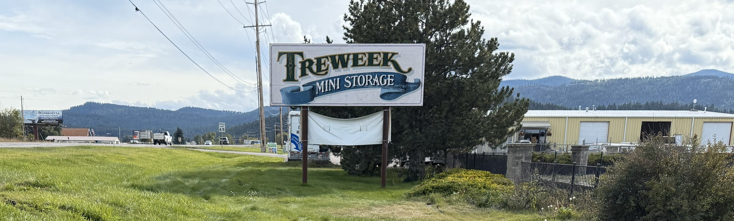 Freeweek Mini Storage sign on grass by a road, with trees, industrial buildings, and mountains in the background.