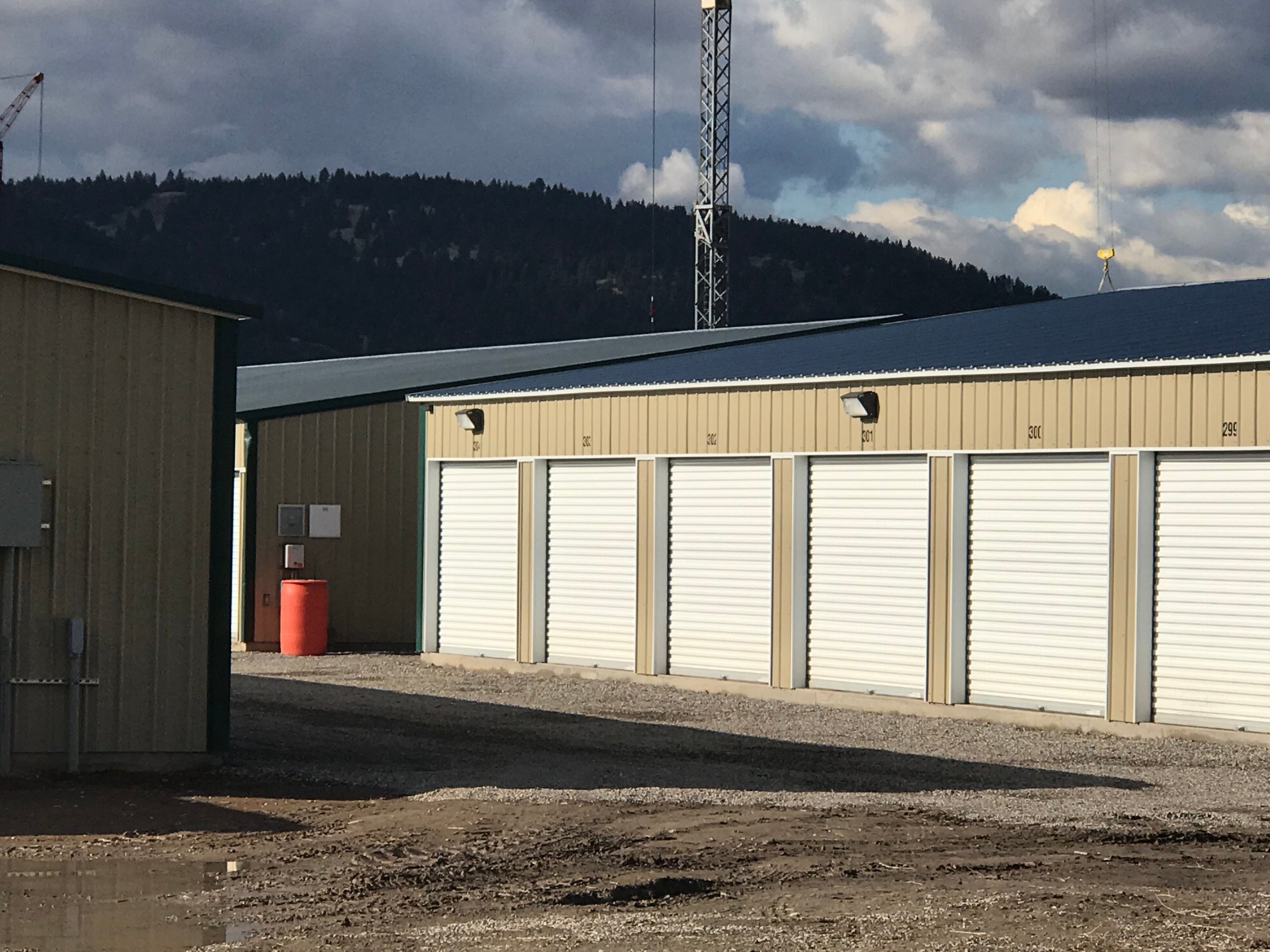 Row of beige storage units with white roll-up doors under a cloudy sky and mountainous background.