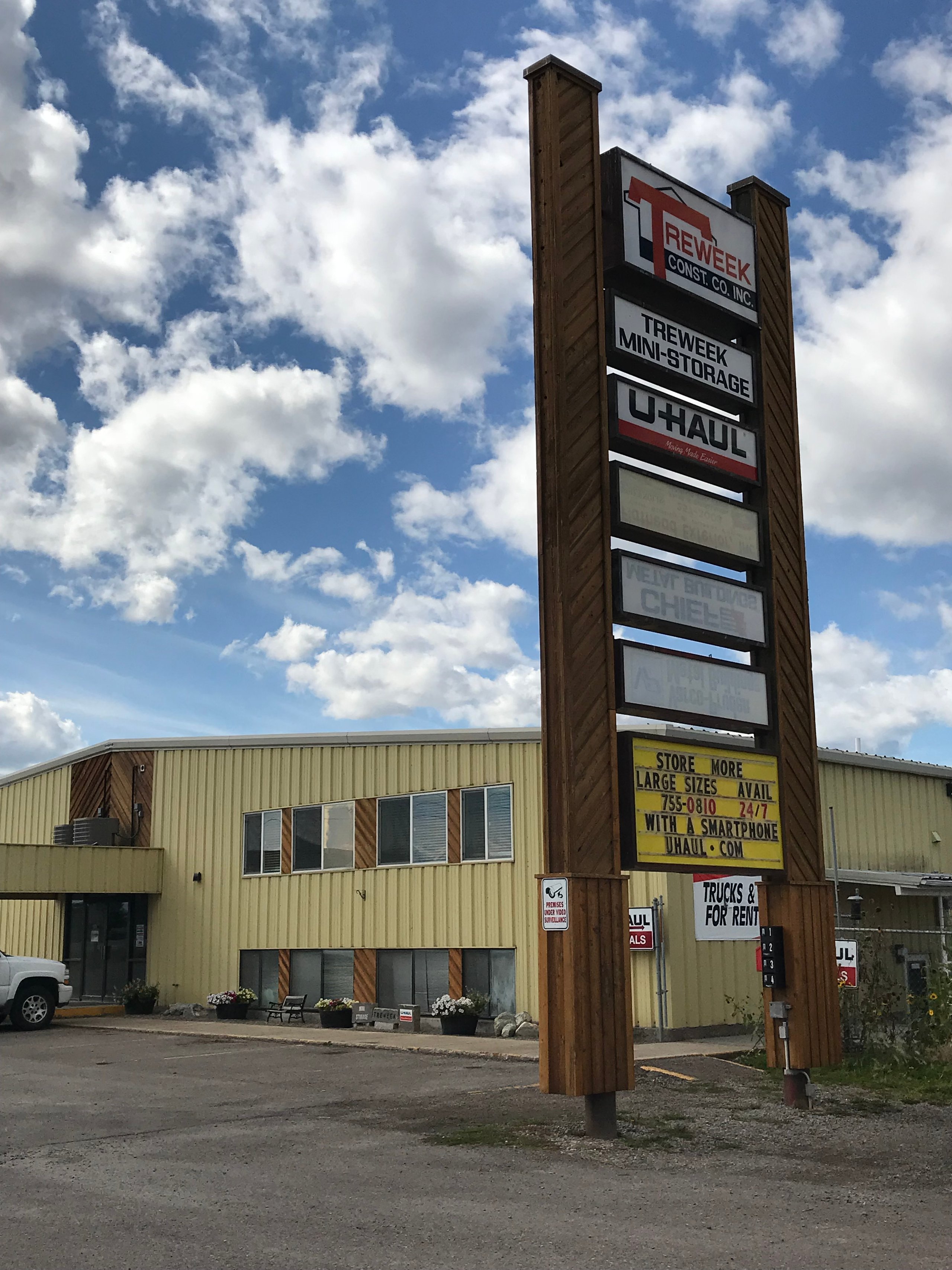 Tall wooden signboard for Treweek Mini Storage and U-Haul in front of a beige industrial building under a partly cloudy sky.