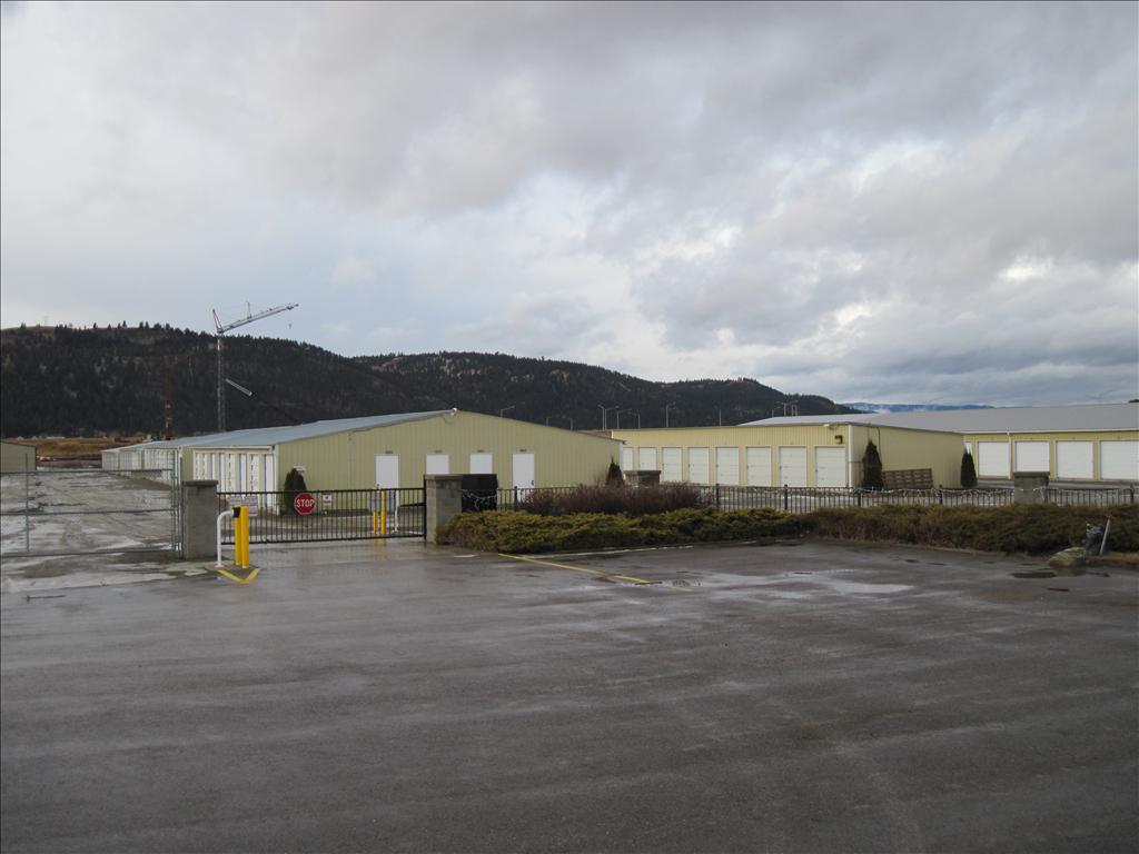 Gated storage facility with rows of yellow storage units and a wet asphalt driveway under a cloudy sky.