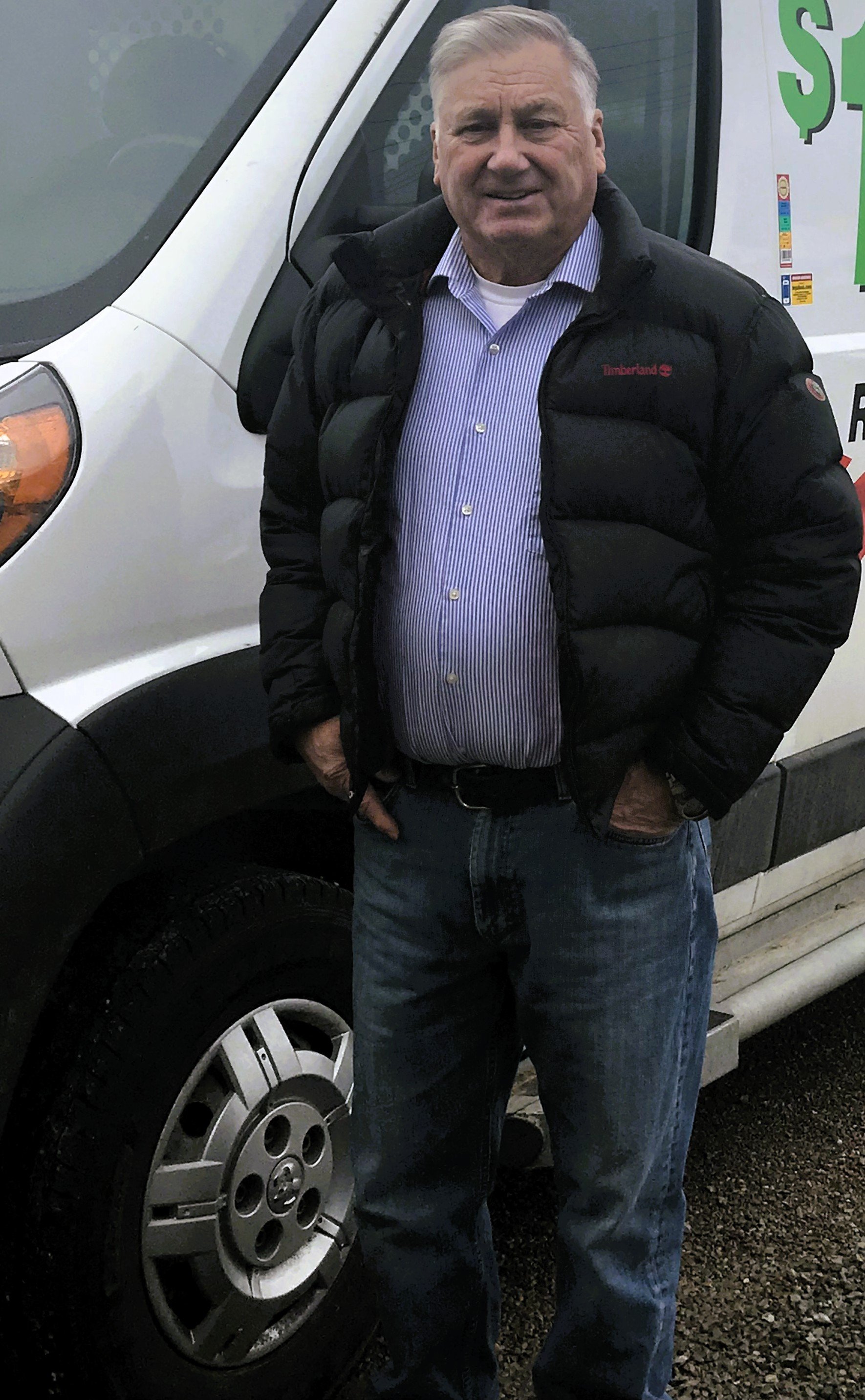 Older man wearing a black Timberland jacket, striped shirt, and jeans standing beside a white van.