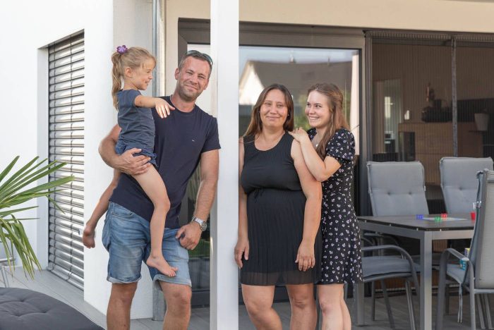 Einfamilienhaus mit Flachdach und großen Glasfronten, überdachte Terrasse und Familie vor dem Eingang
