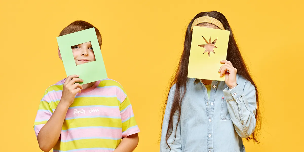 Two children holding colored paper with cutouts over their faces against a yellow background, one with a square cutout and the other with a star-shaped cutout.