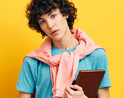 Young person with curly hair wearing a blue t-shirt and a pink sweatshirt draped over shoulders holding a brown notebook against a yellow background.