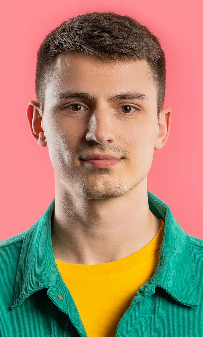Young man with short brown hair wearing a green jacket and yellow shirt against a pink background.