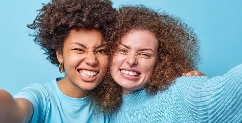 Two young women with curly hair smiling and taking a selfie against a light blue background.