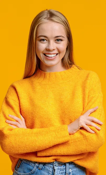 Smiling young woman with long blonde hair wearing an orange sweater and blue jeans, standing with arms crossed against an orange background.
