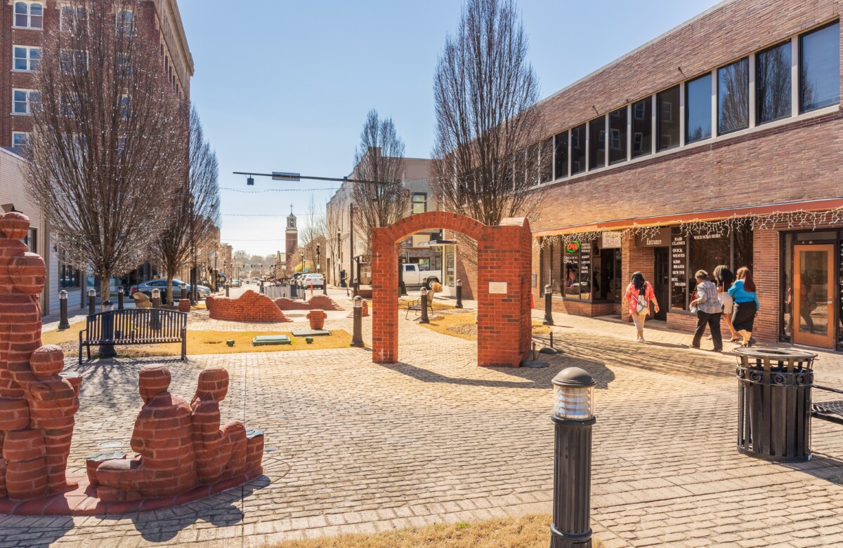 A sunny pedestrian plaza in a small downtown area with brick paving, red brick archway and sculptures, leafless trees, benches, and storefronts along the street; a few people walk past shops on the right while cars are visible in the distance.