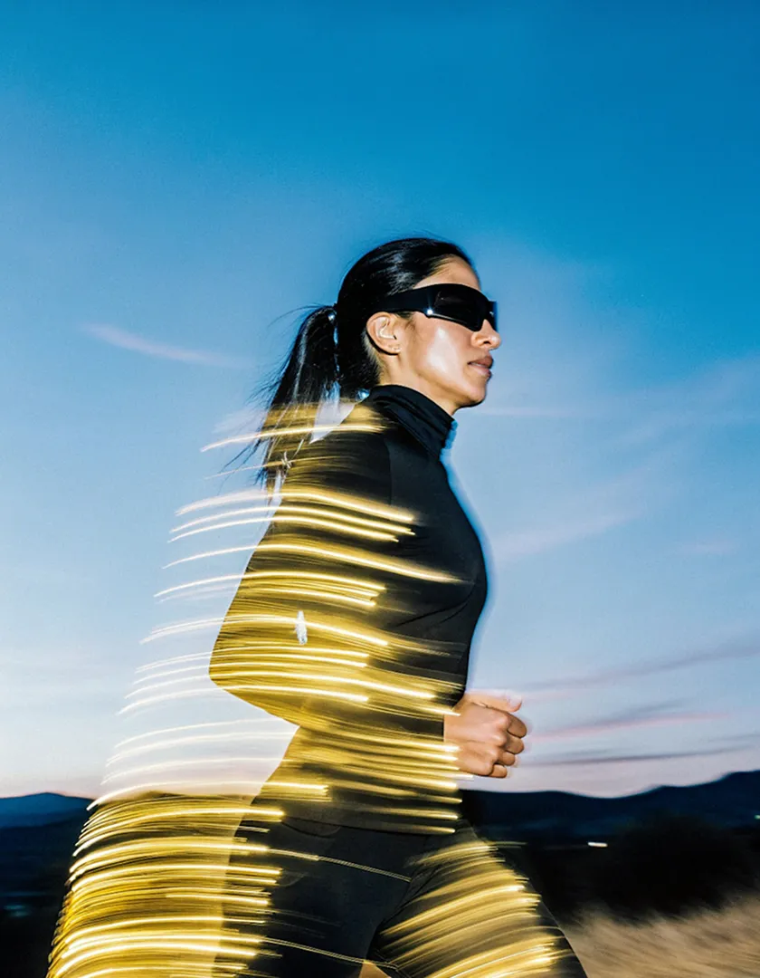 Woman in black athletic wear and sunglasses running outdoors at dusk with light trails wrapped around her.
