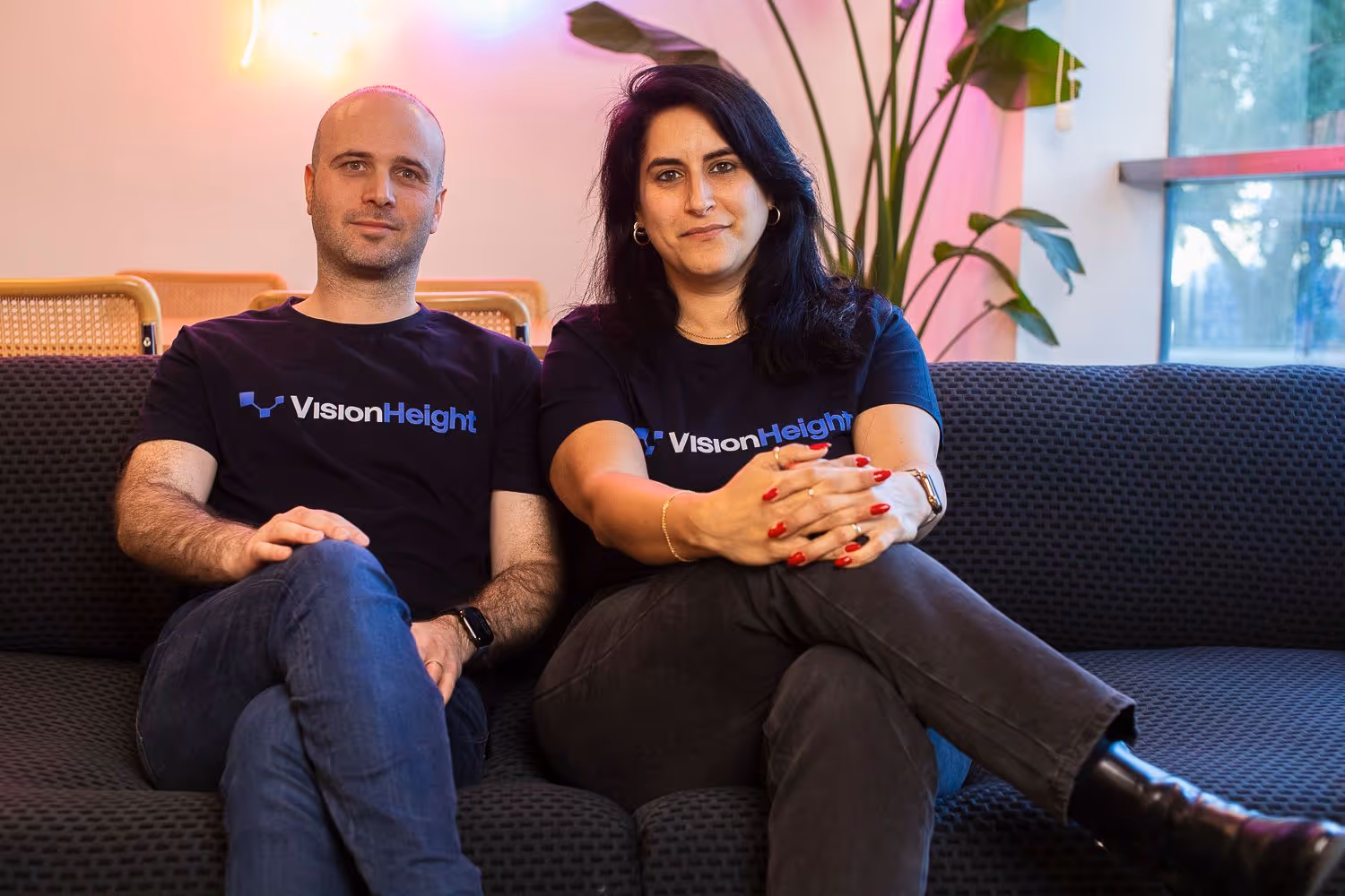 Smiling man and woman sitting side by side on a couch wearing VisionHeight t-shirts.
