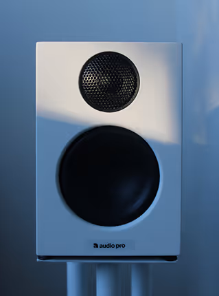 White Audio Pro speaker with a black tweeter and woofer on a stand against a blurred blue background.