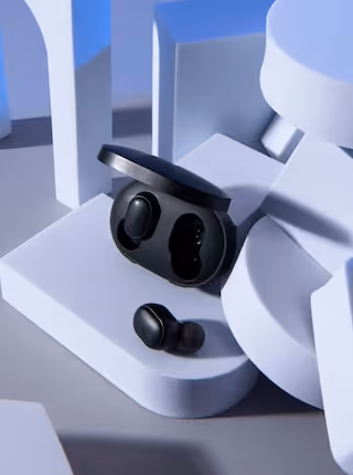 Black wireless earbuds with one earbud outside and the other inside an open charging case placed on white geometric shapes.