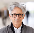 Older man with gray hair and glasses wearing a dark jacket and white shirt, smiling slightly.