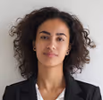 Woman with curly hair wearing a black blazer and white top, looking at the camera with a neutral expression.
