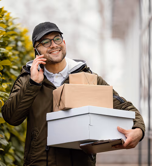 Smiling delivery man wearing glasses and a cap, holding multiple packages and talking on a cellphone outdoors.