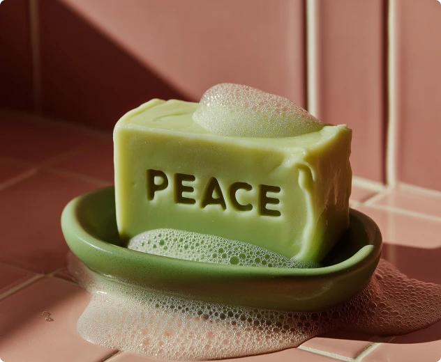 Green soap bar carved with the word PEACE sitting on a green dish with soap suds over pink tiles.