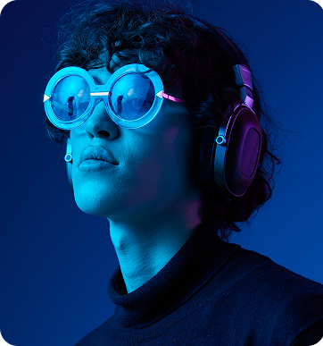 Person with curly hair wearing large round glasses and over-ear headphones, illuminated by blue light against a dark background.