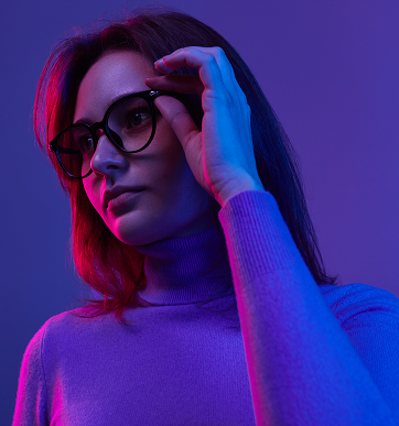 Woman wearing glasses and a purple turtleneck illuminated by pink and blue lighting against a dark background.