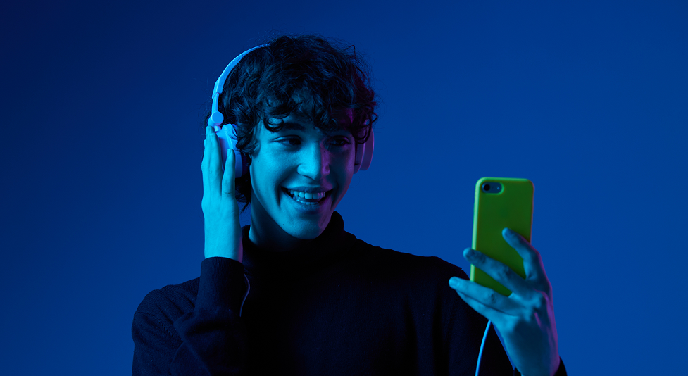 Young man with curly hair wearing white headphones smiling while holding a yellow smartphone against a blue background.