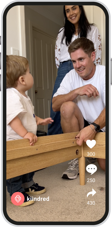 Man crouching and smiling at toddler playing with wooden furniture, woman standing smiling behind them.