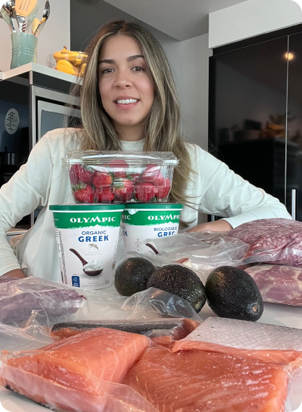 Woman smiling behind a kitchen counter displaying packaged fresh salmon, avocados, Greek yogurt, strawberries, and cuts of meat.