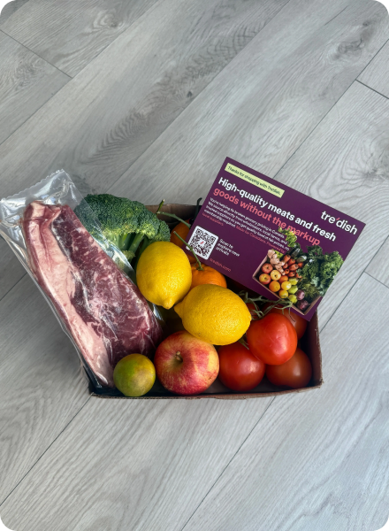 Box containing packaged raw beef, broccoli, apples, lemons, tomatoes, and a promotional card on light wooden floor.
