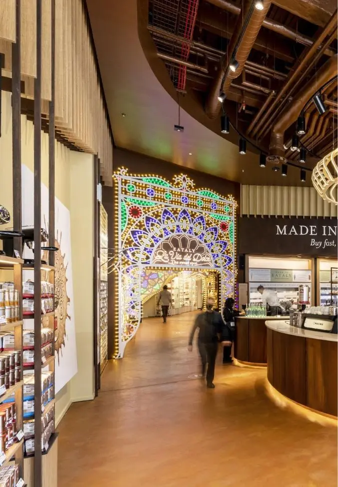 Interior of a modern marketplace with a colorful, illuminated archway labeled 'EATALY Via del Dolo' leading to a brightly lit corridor, people walking, and wooden display shelves and counters.
