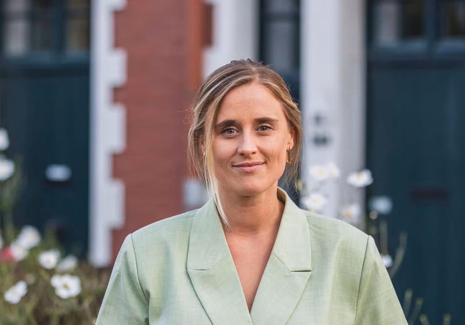 Portrait of a woman with blonde hair wearing a light green blazer standing outdoors with blurred buildings and white flowers in the background.
