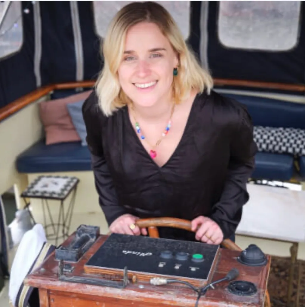 Vrouw met blond haar en zwarte blouse glimlacht terwijl ze een houten stuur vasthoudt op een boot.