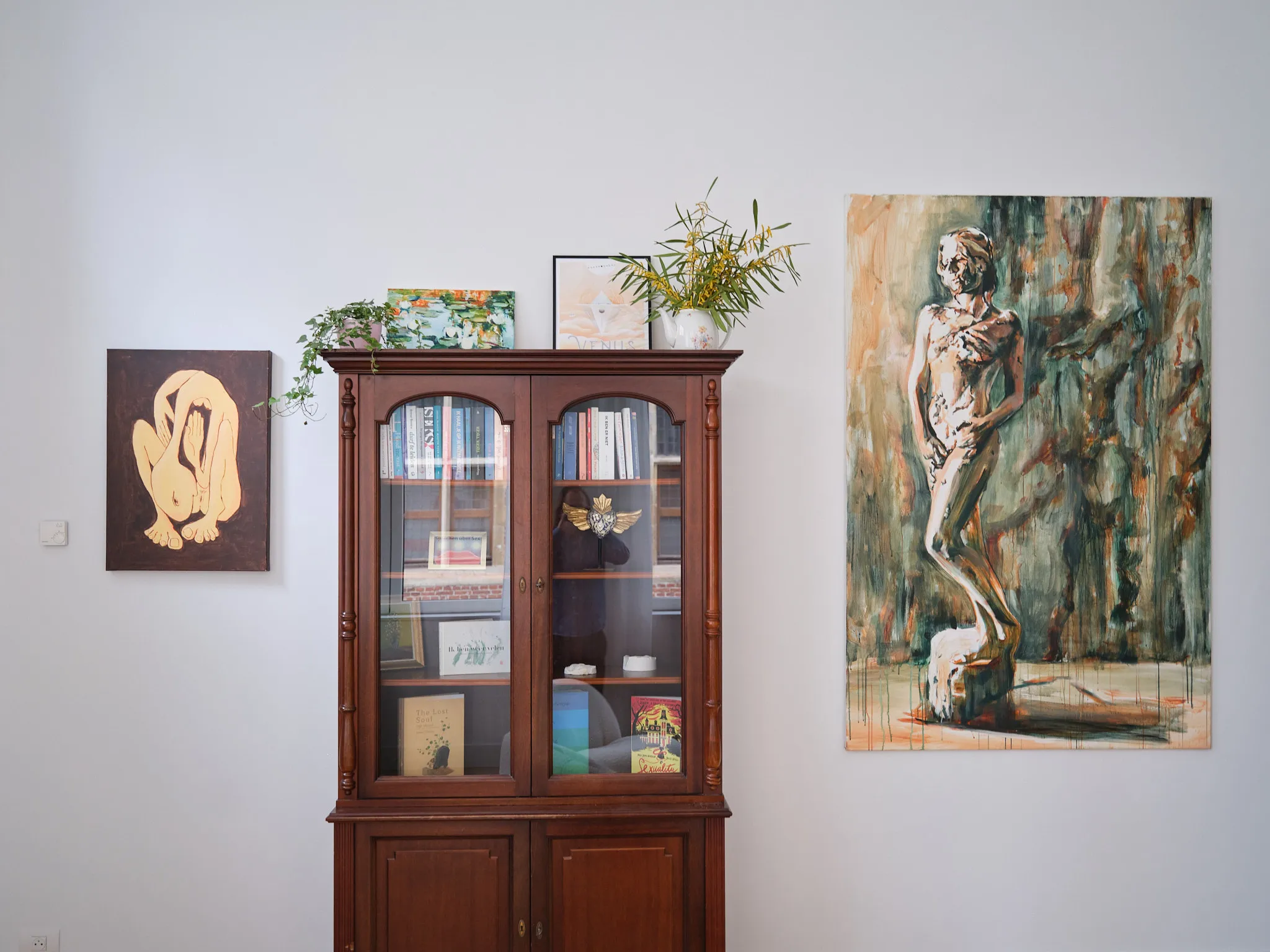 Houten boekenkast met glazen deuren gevuld met boeken en een decoratief item, geflankeerd door twee schilderijen aan de muur en twee schilderijen en een vaas met planten op de kast.