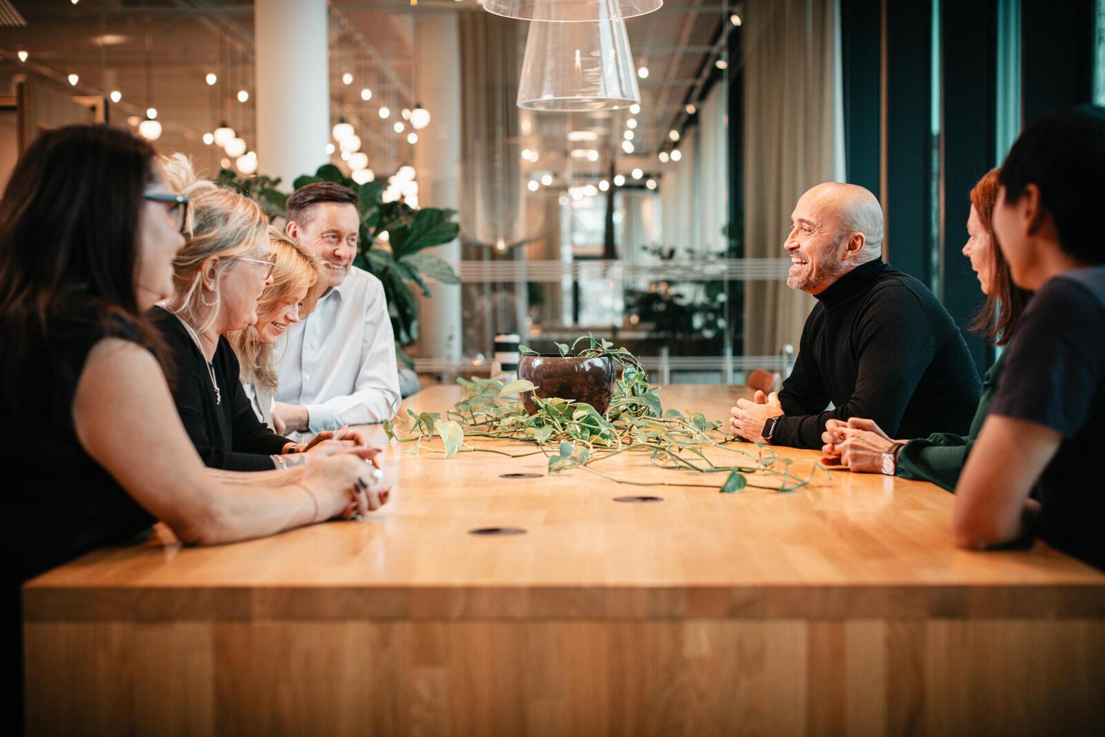 Grupp av glada personer sitter runt ett stort träbord i ett modernt mötesrum med växt i mitten.