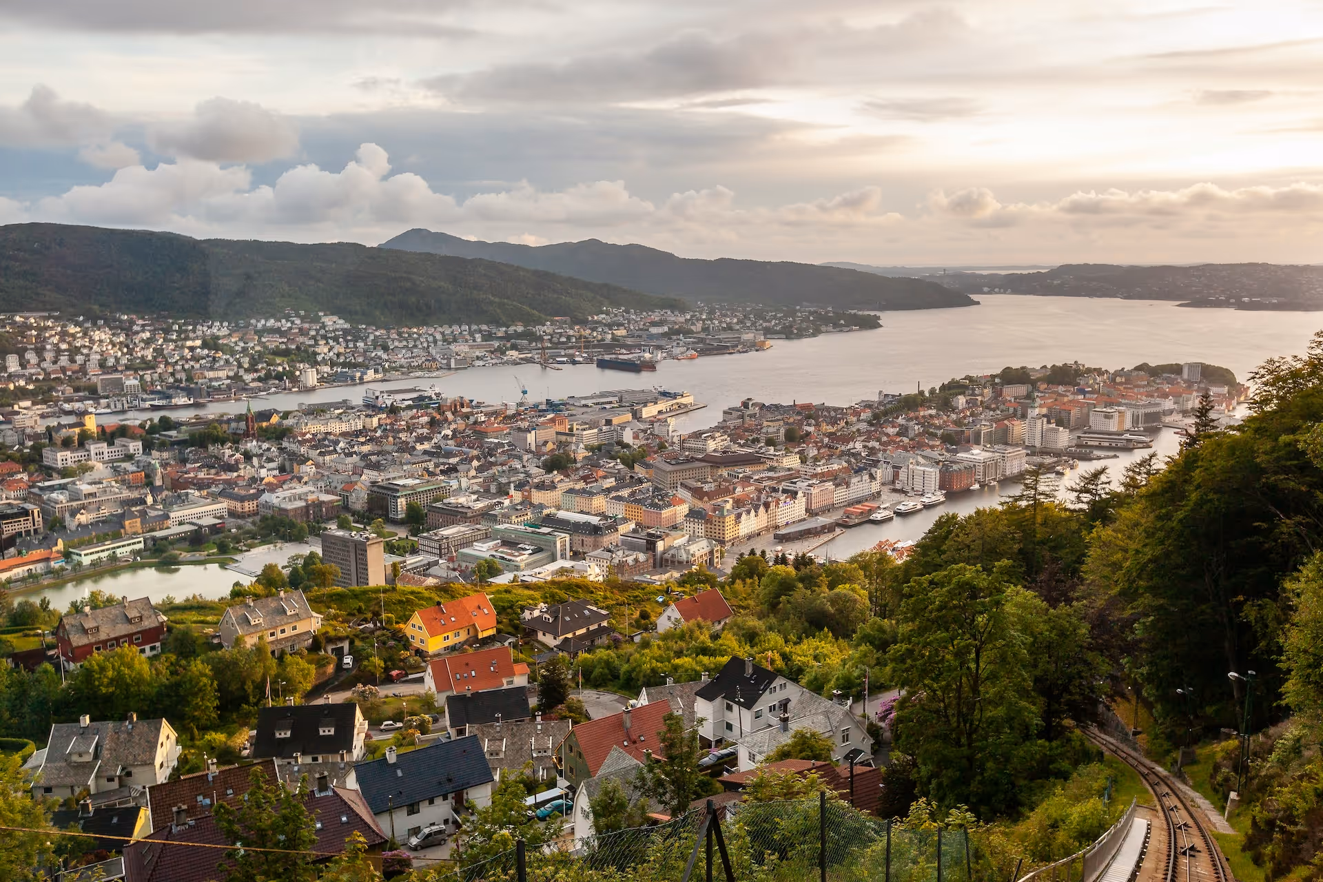 Oversiktsbilde av Bergen by ved vannet med fargerike hus, grønne trær og fjell i bakgrunnen ved solnedgang.
