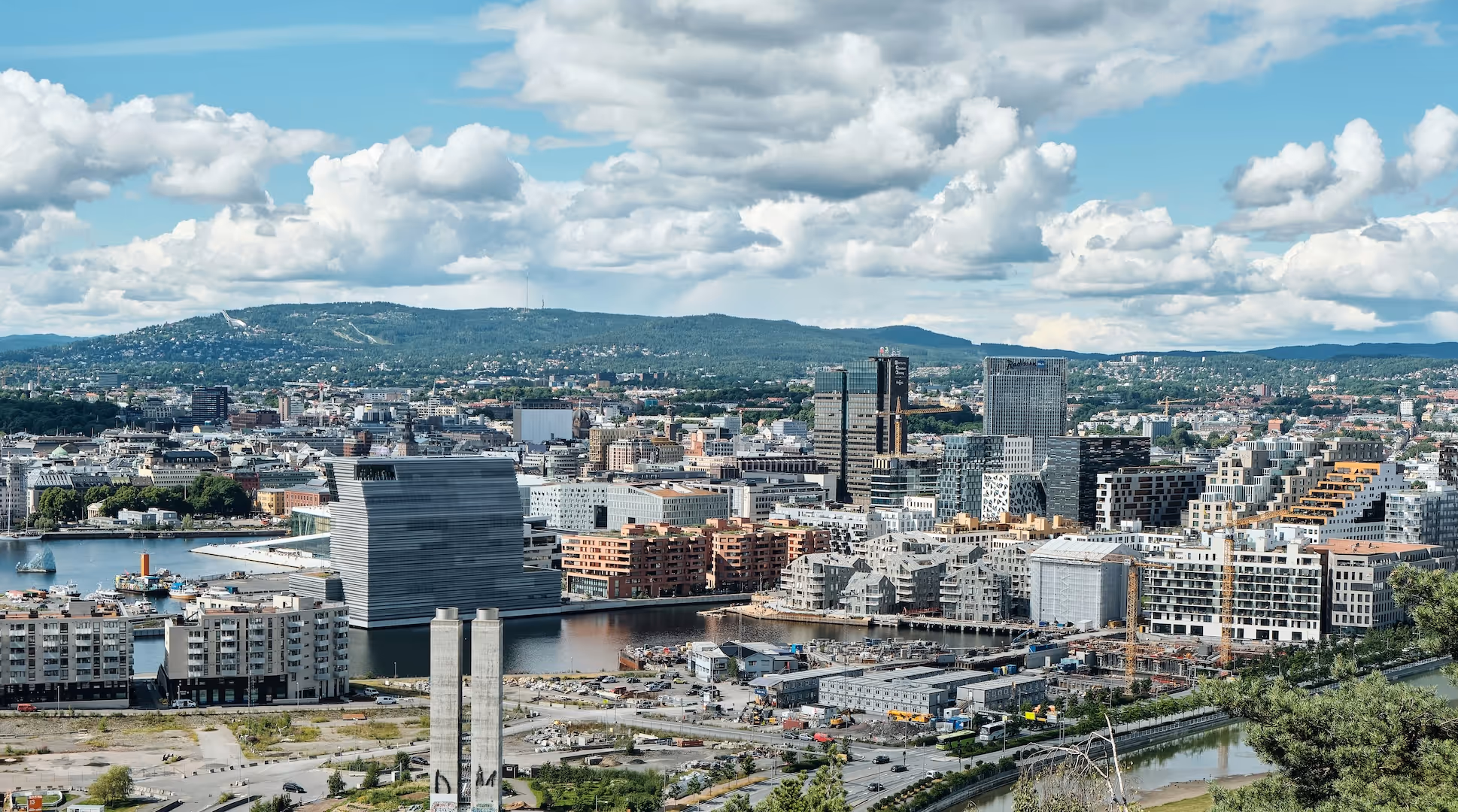 Utsikt over Oslo by med moderne bygninger, havn og grønne åser i bakgrunnen under en blå himmel med skyer.