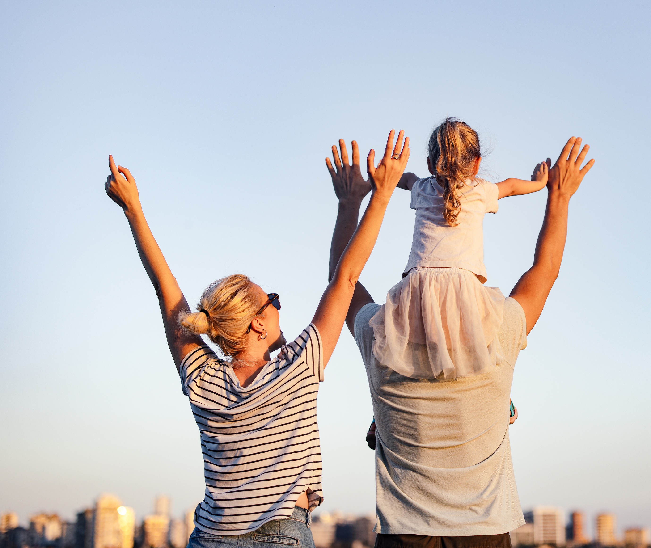 Familie med mor, far og datter vendt vekk fra kamera og holder hendene oppe i luften. Belyst av sol bakfra med blå himmel foran.