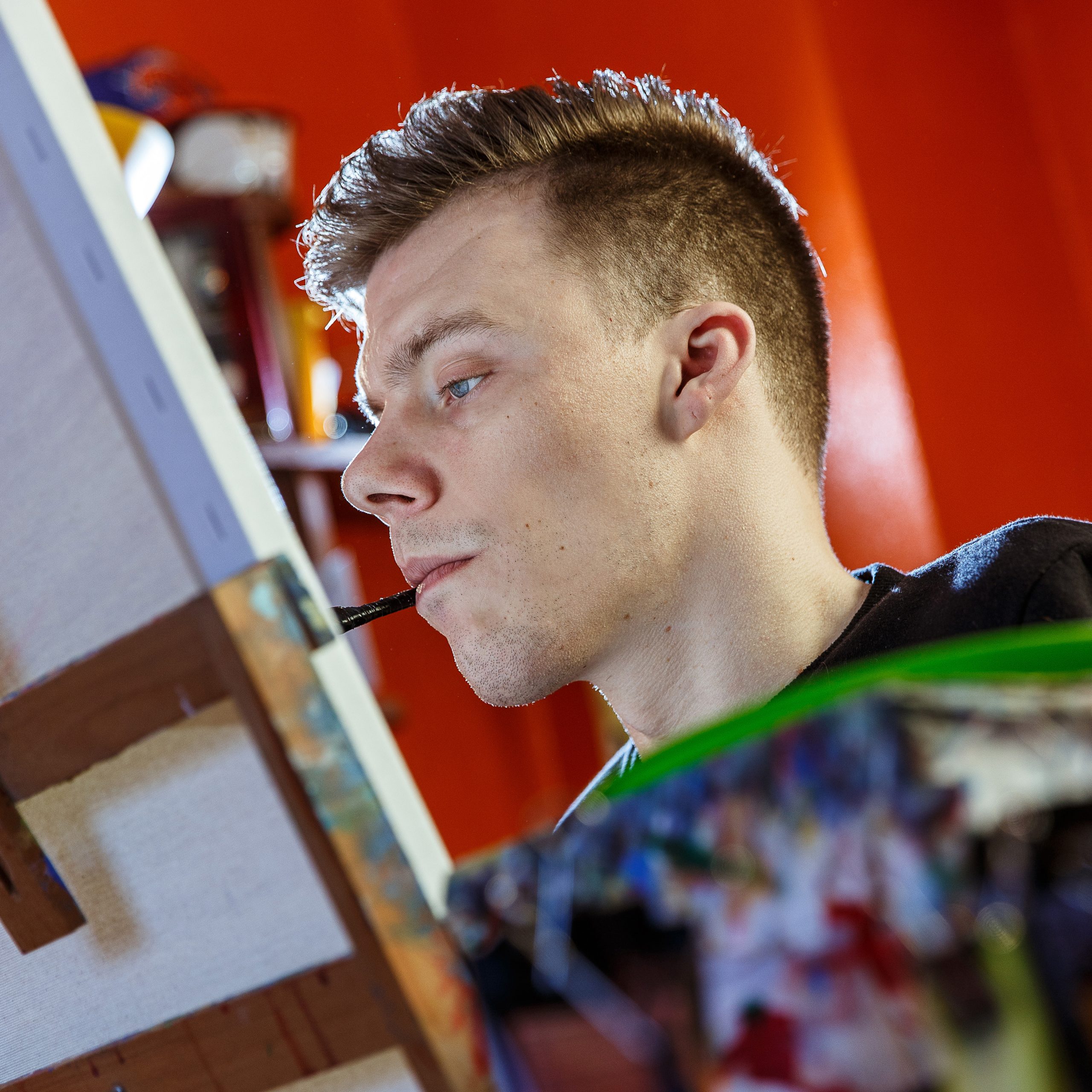 Young man concentrating while holding a paintbrush in his mouth, painting on a canvas.