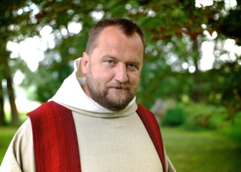 Man wearing a beige robe with red vertical stripes standing outdoors with trees in the background.