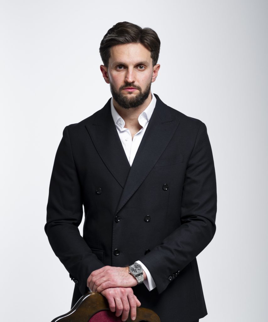 Serious man with dark hair and beard wearing a black double-breasted blazer and white shirt, resting hands on the back of a chair.