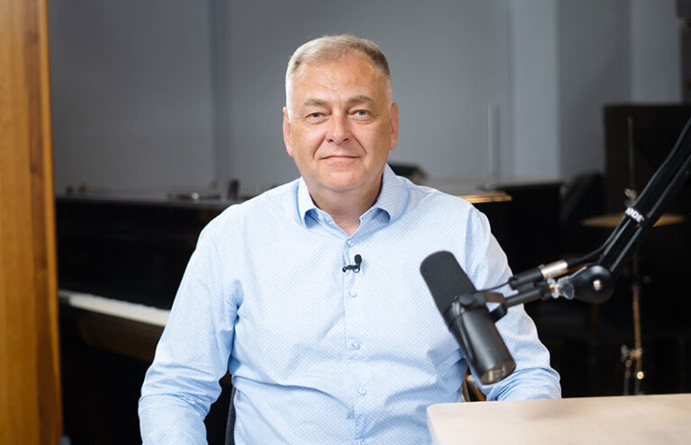 A middle-aged man in a light blue shirt sitting in front of a microphone and smiling, with a piano in the background.