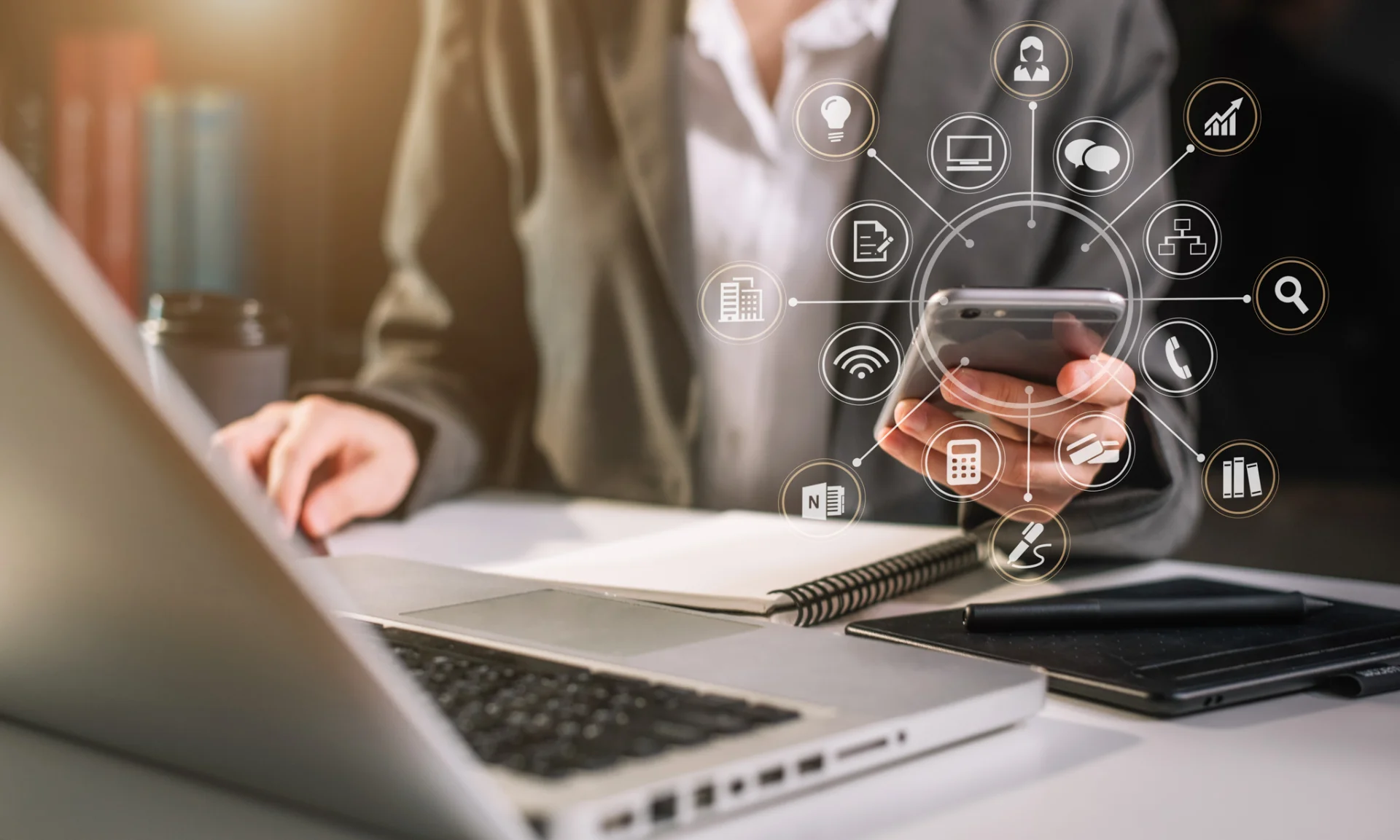 Person hält Smartphone mit überlagerten Icons für Kommunikation und Datenmanagement neben offenem Laptop und Notizbuch.