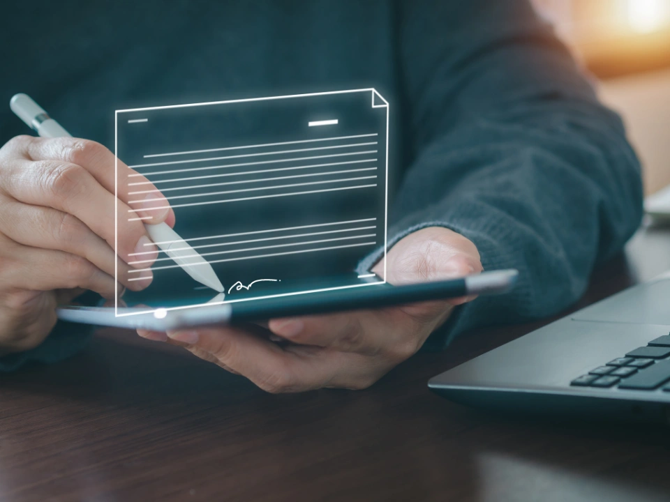 Person unterschreibt digitales Dokument mit einem Stift auf einem Tablet neben einem Laptop.