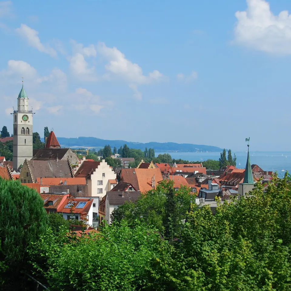 Blick auf eine Stadt mit roten Dächern, Kirchtürmen und grünen Bäumen im Vordergrund, im Hintergrund ein großer See unter blauem Himmel.