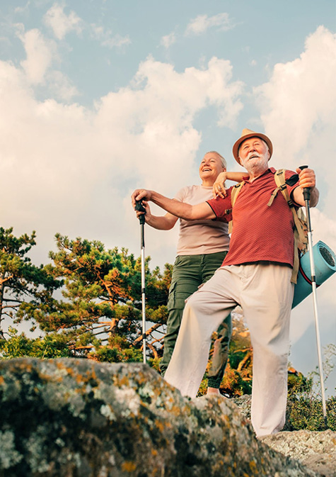 Smiling elderly couple hiking outdoors using walking poles, surrounded by trees and rocks under a partly cloudy sky.